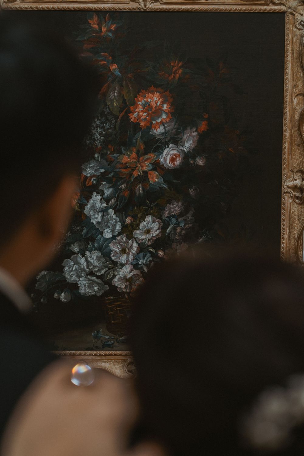 Couple admiring a classic floral still life painting in an ornate gold frame at an art museum.