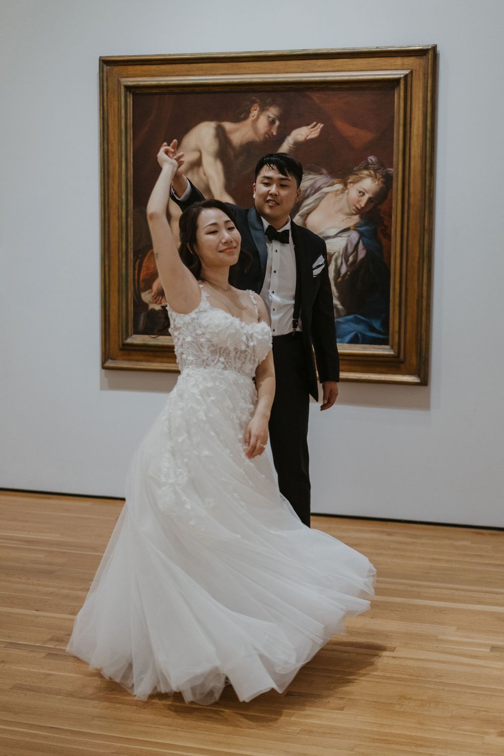 Bride and groom dancing joyfully in front of a classical painting at an art museum wedding.