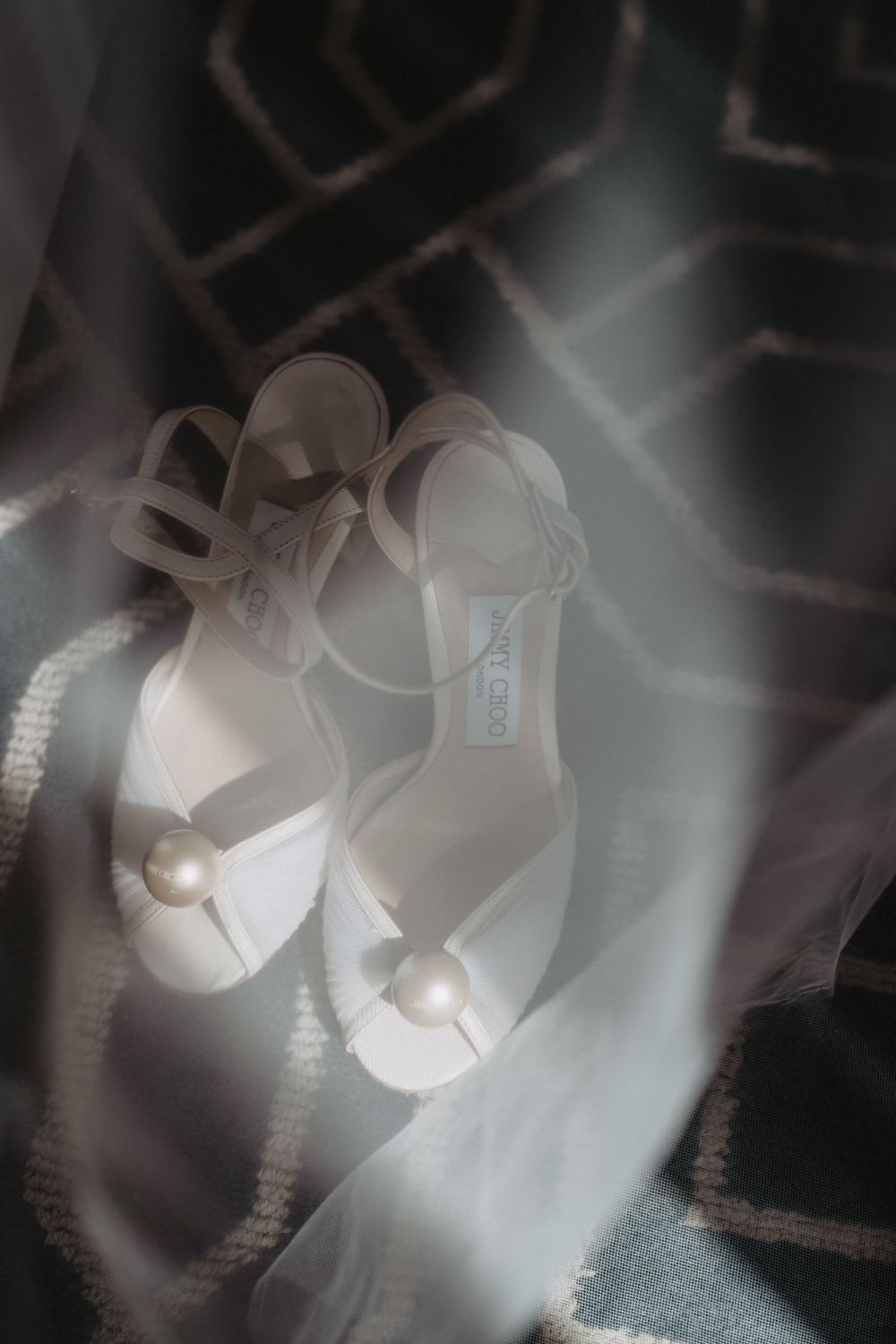 Elegant white Jimmy Choo bridal heels with pearl embellishments resting on a patterned surface in soft light.