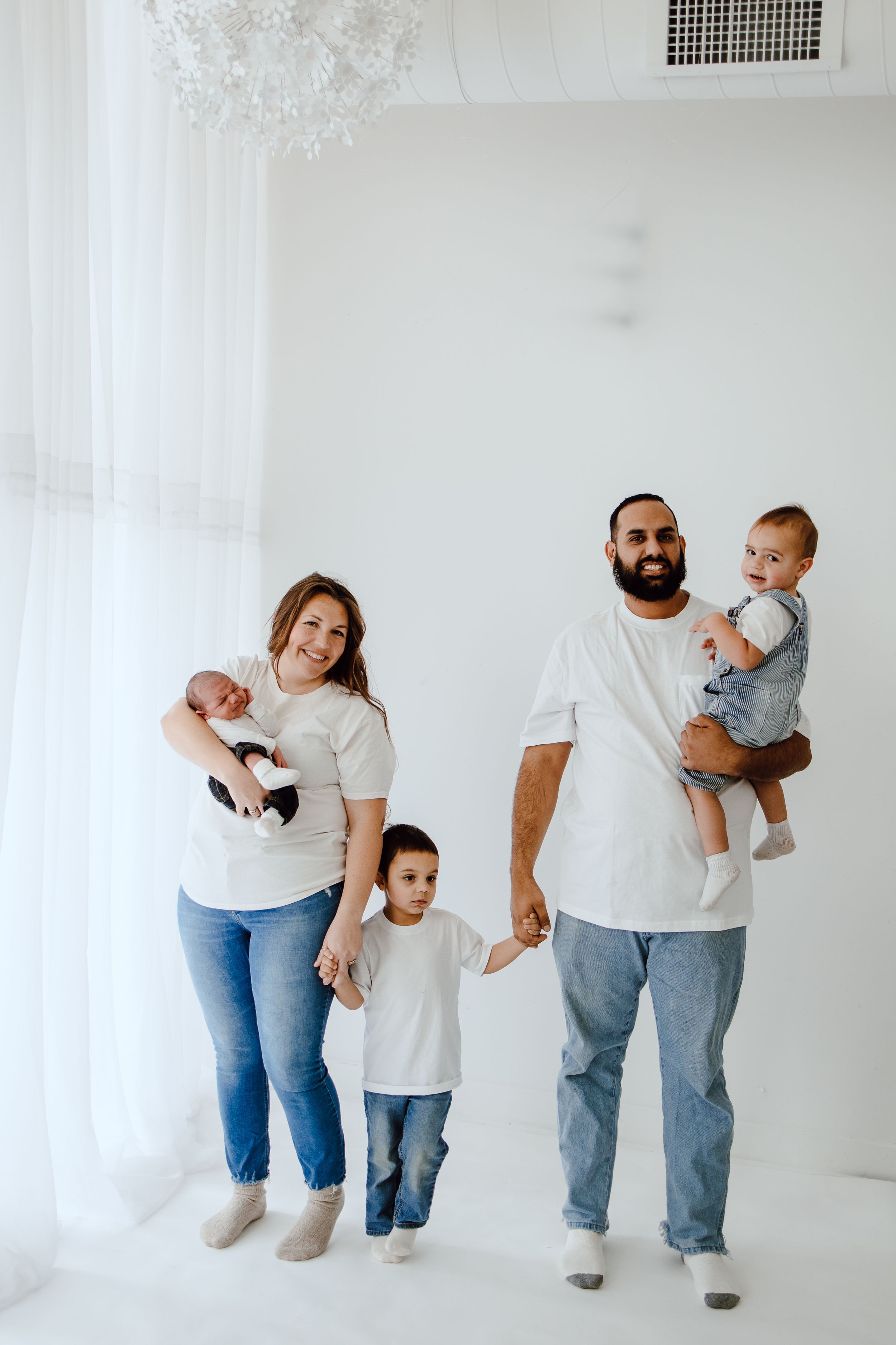 Family wearing matching white t-shirts and jeans poses together against white wall in minimalist studio setting.