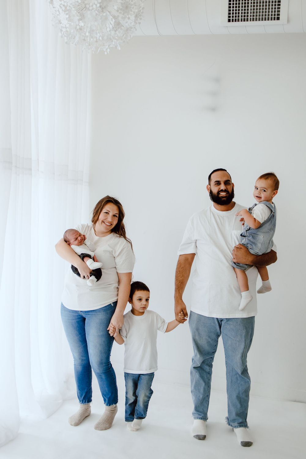 Family wearing matching white t-shirts and jeans poses together against white wall in minimalist studio setting.