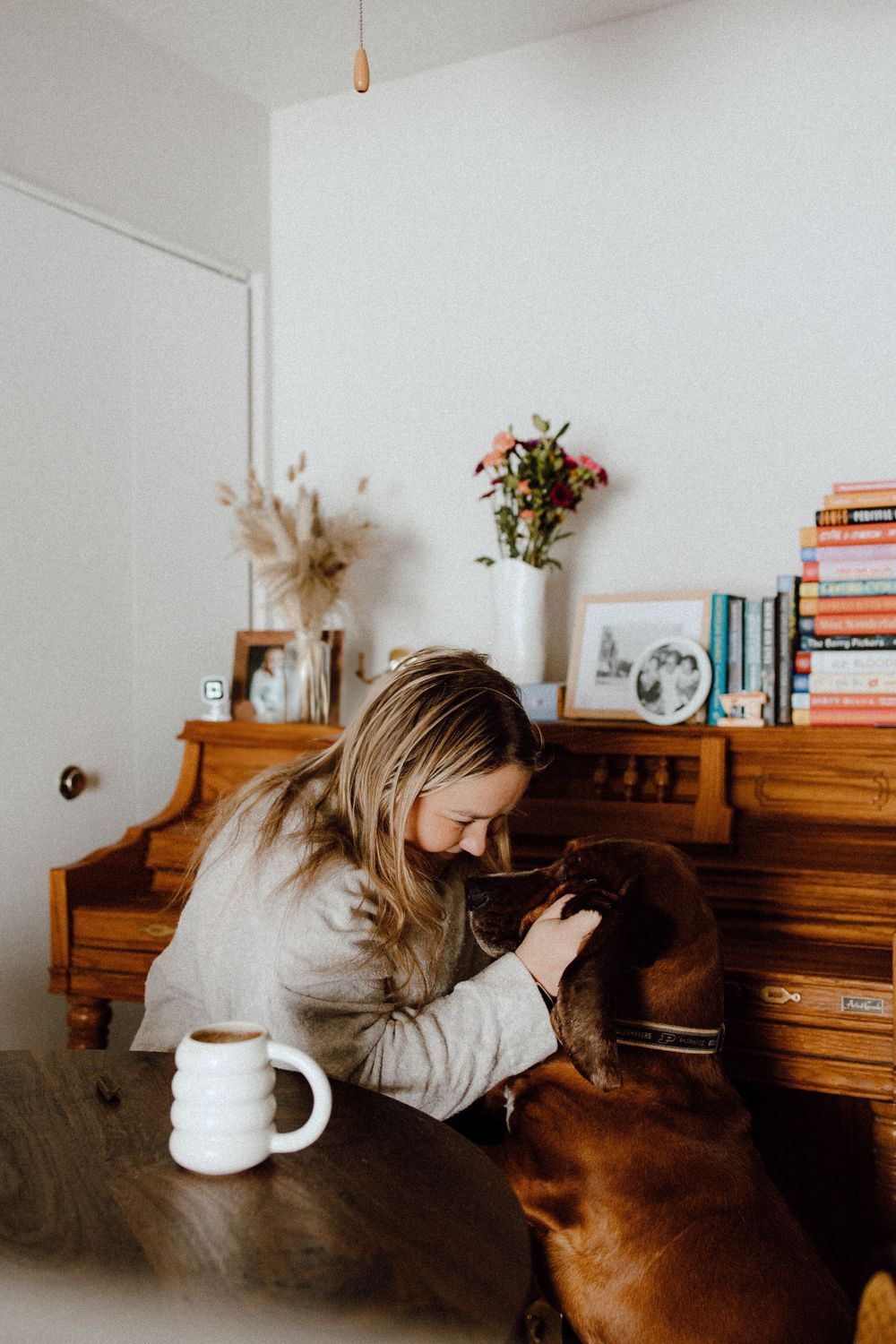 A cozy home scene with a brown dog and person sitting by a wooden piano adorned with books and flowers.