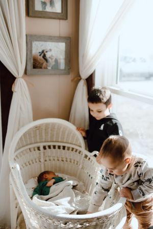 Two children stand beside a white wicker bassinet near a window with flowing curtains in a softly lit nursery interior.