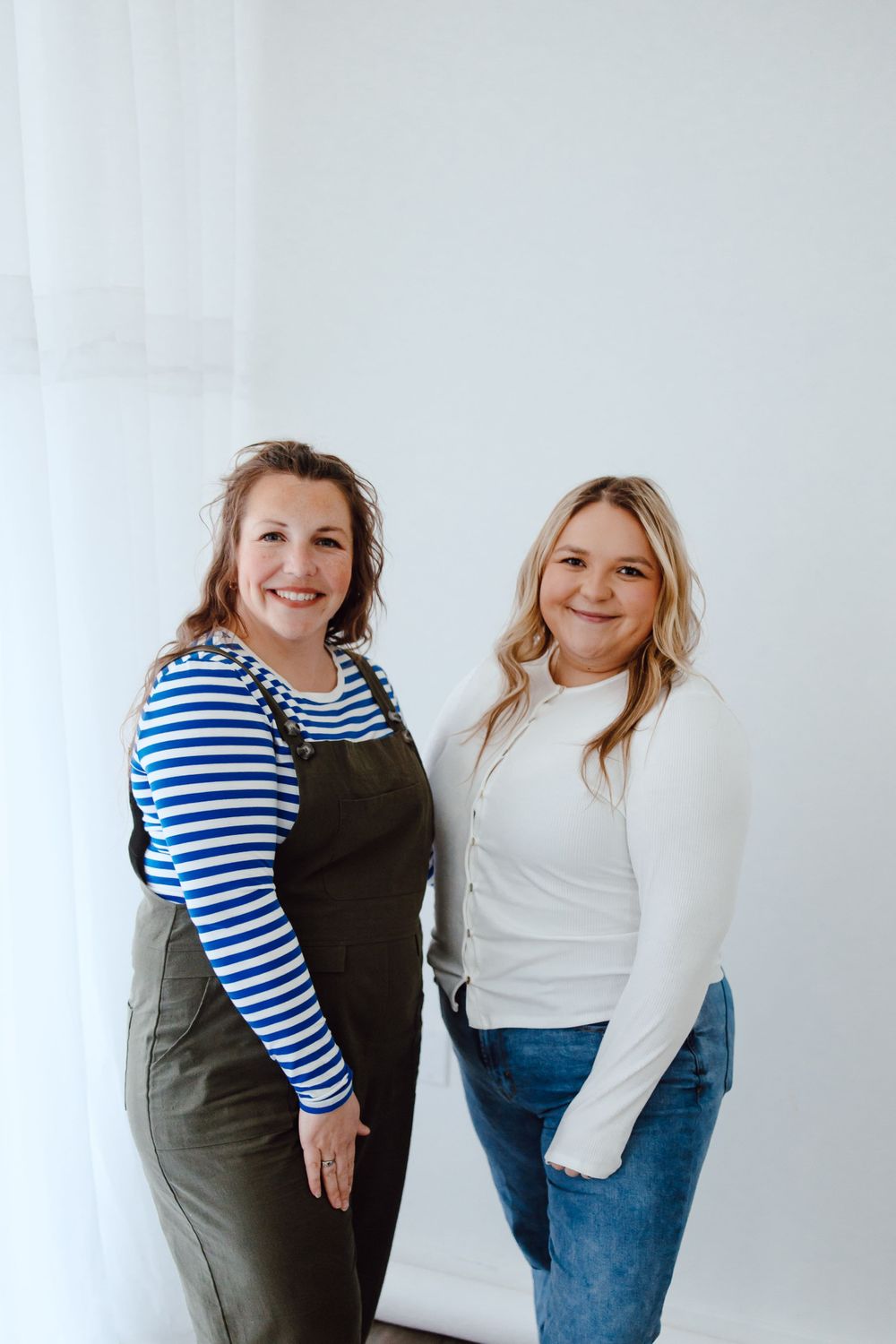 Two people stand together in casual clothing, wearing a striped top and overalls and a white sweater against a white backdrop.