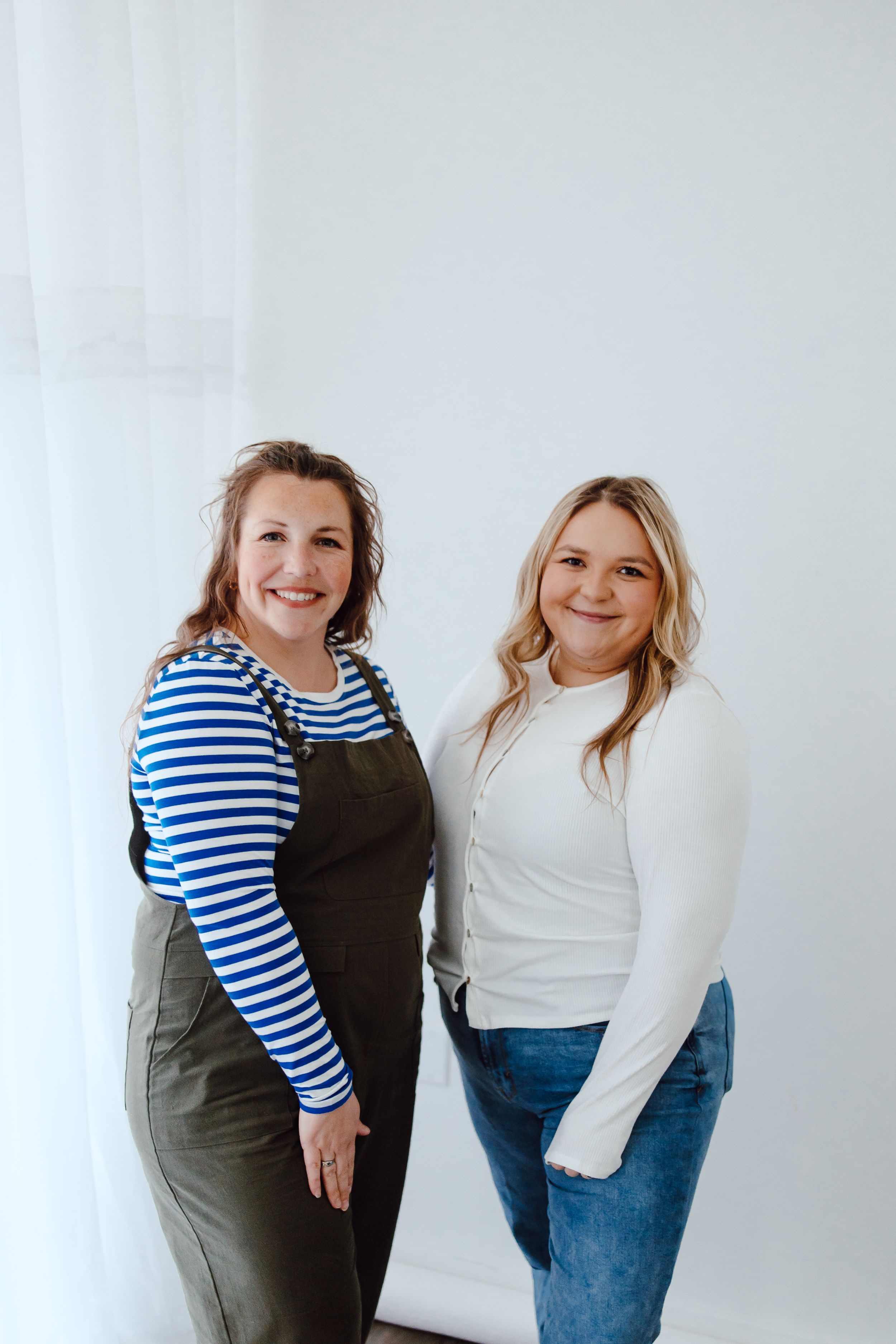Two people stand together in casual clothing, wearing a striped top and overalls and a white sweater against a white backdrop.