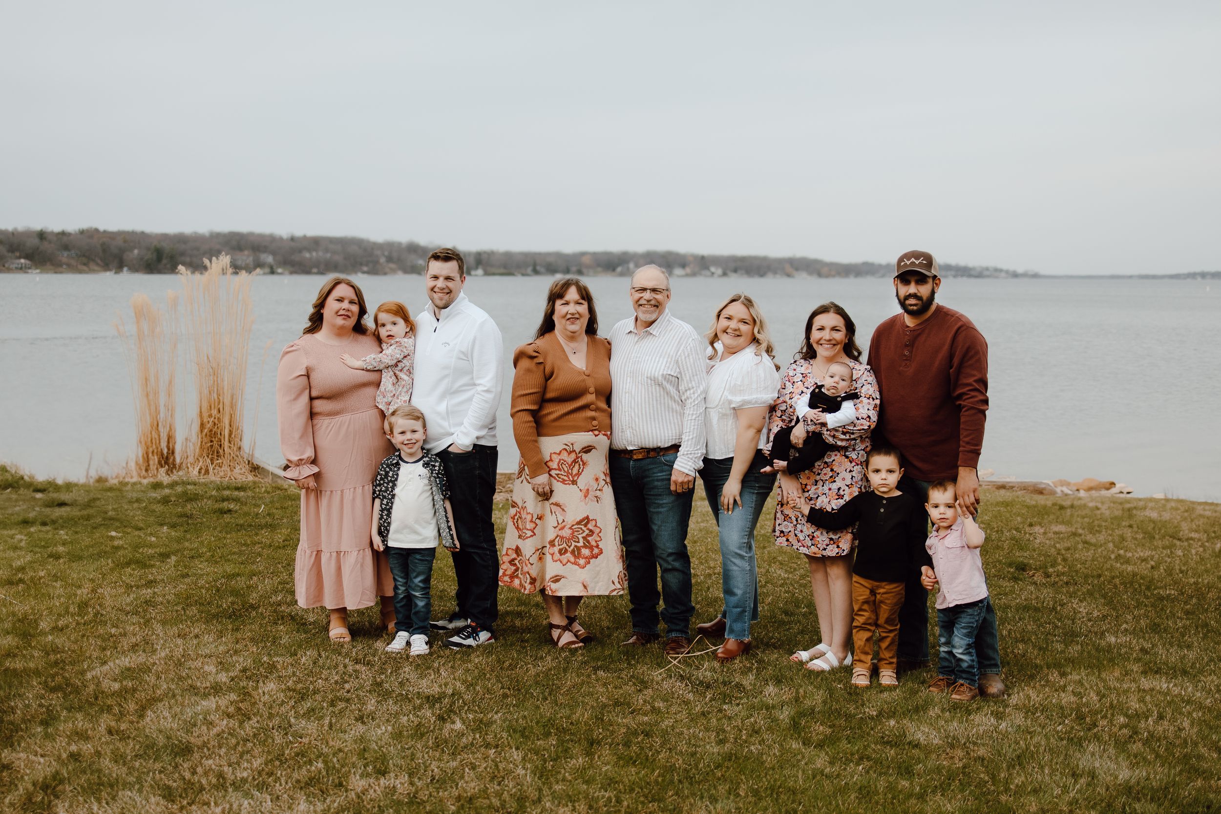 Group gathered on grassy hill overlooking water on cloudy day wearing coordinated autumn colors and casual attire.