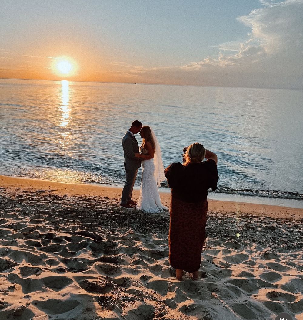 A photographer captures a couple's silhouette on a sandy beach during a stunning golden hour sunset over calm waters.