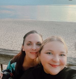 Two friends taking a selfie together at the beach during sunset with calm waters in the background.
