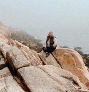 Person in warm clothing sits on large granite rocks along misty coastal cliffs.