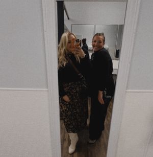 Two people in casual outfits laughing together while taking a mirror selfie in what appears to be a hallway.