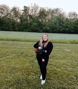 Someone standing in a grassy field wearing all black clothing against a backdrop of trees at dusk.