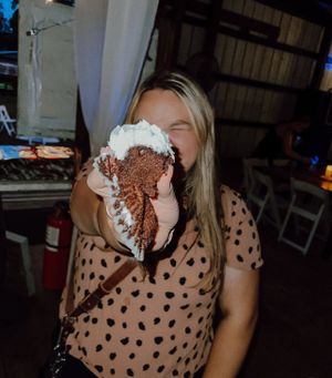 Someone in a polka dot top enthusiastically bites into a messy dessert covered in whipped cream and chocolate.