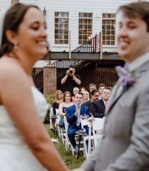 Wedding guests sitting in rows of white chairs watch a couple's ceremony at an outdoor venue with brick building in background.