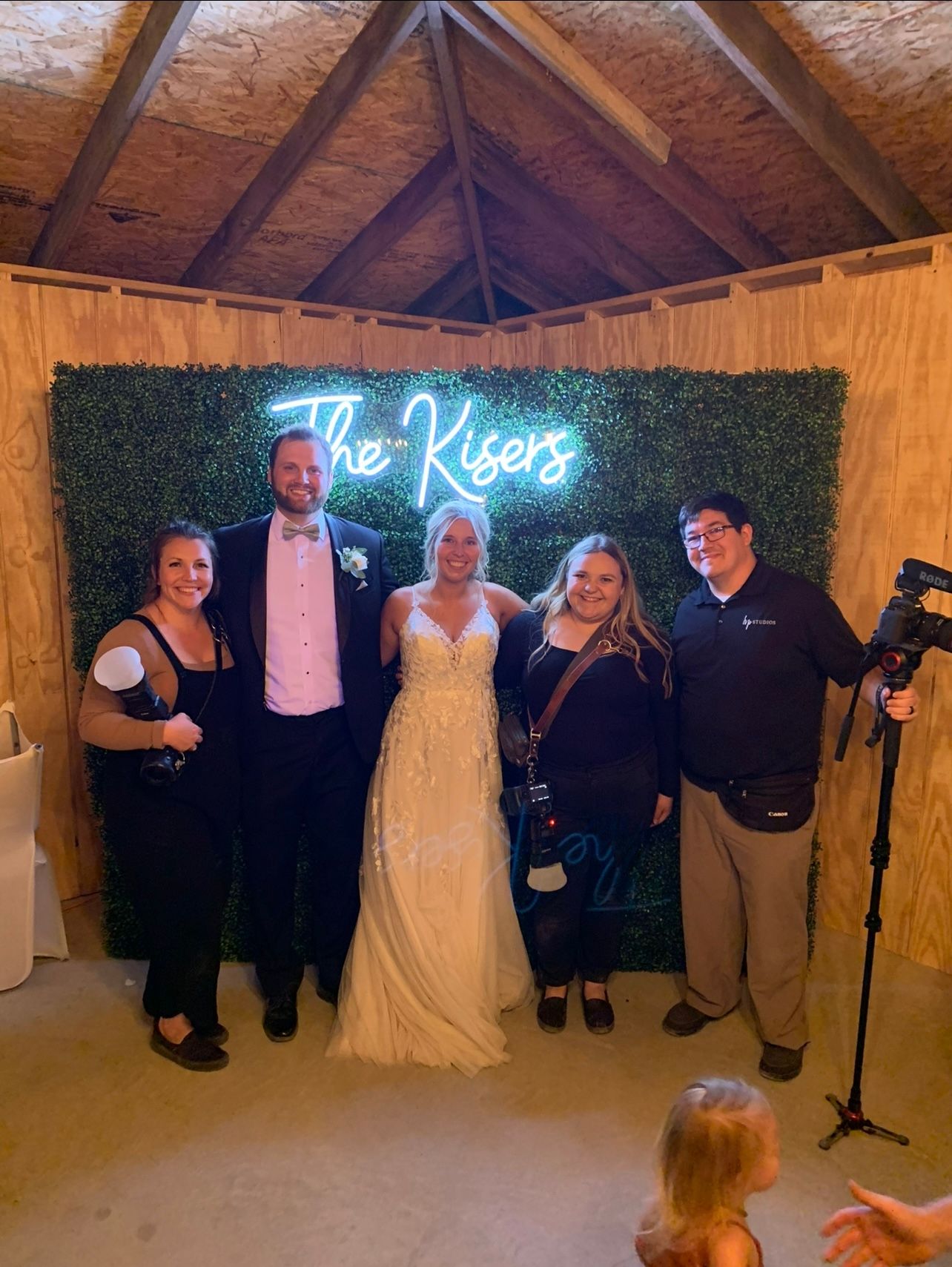 Wedding party posing in front of neon sign and hedge wall backdrop during indoor reception.