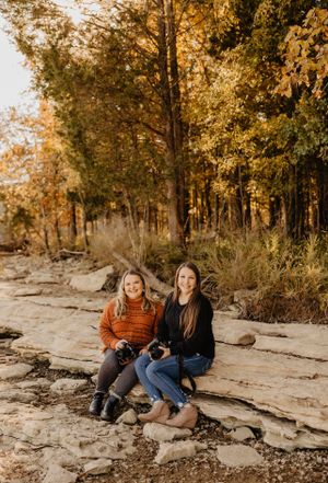Two people sit on rocky ledge surrounded by golden autumn trees in warm sunset lighting.