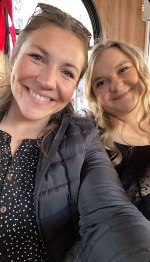 Two smiling people taking a selfie together wearing a polka dot top and black clothing indoors.