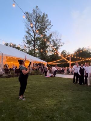 Outdoor evening wedding reception under a white tent with string lights and people gathered on a grassy lawn at dusk.
