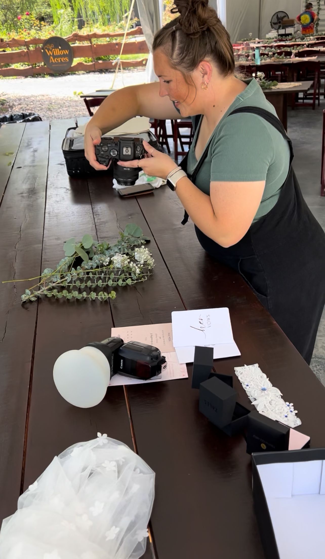 Someone in a sage green shirt takes photos during a setup process with camera equipment and styling supplies on a wooden table.