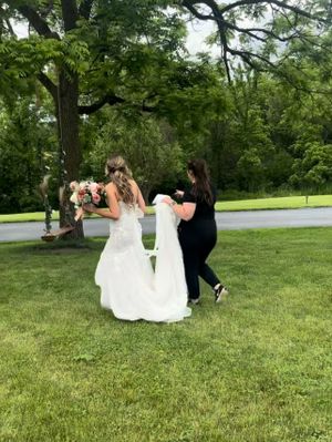 A wedding dress being adjusted outdoors on a grassy lawn surrounded by lush green trees on a summer day.