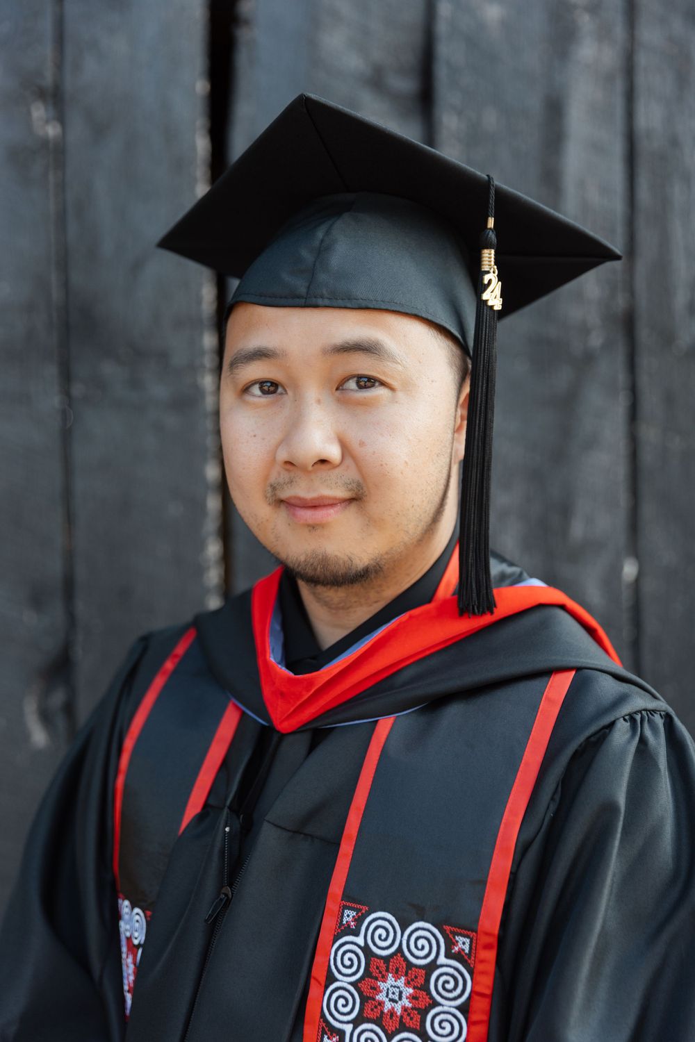Graduate in black academic regalia with red hood and decorative sash poses outdoors.