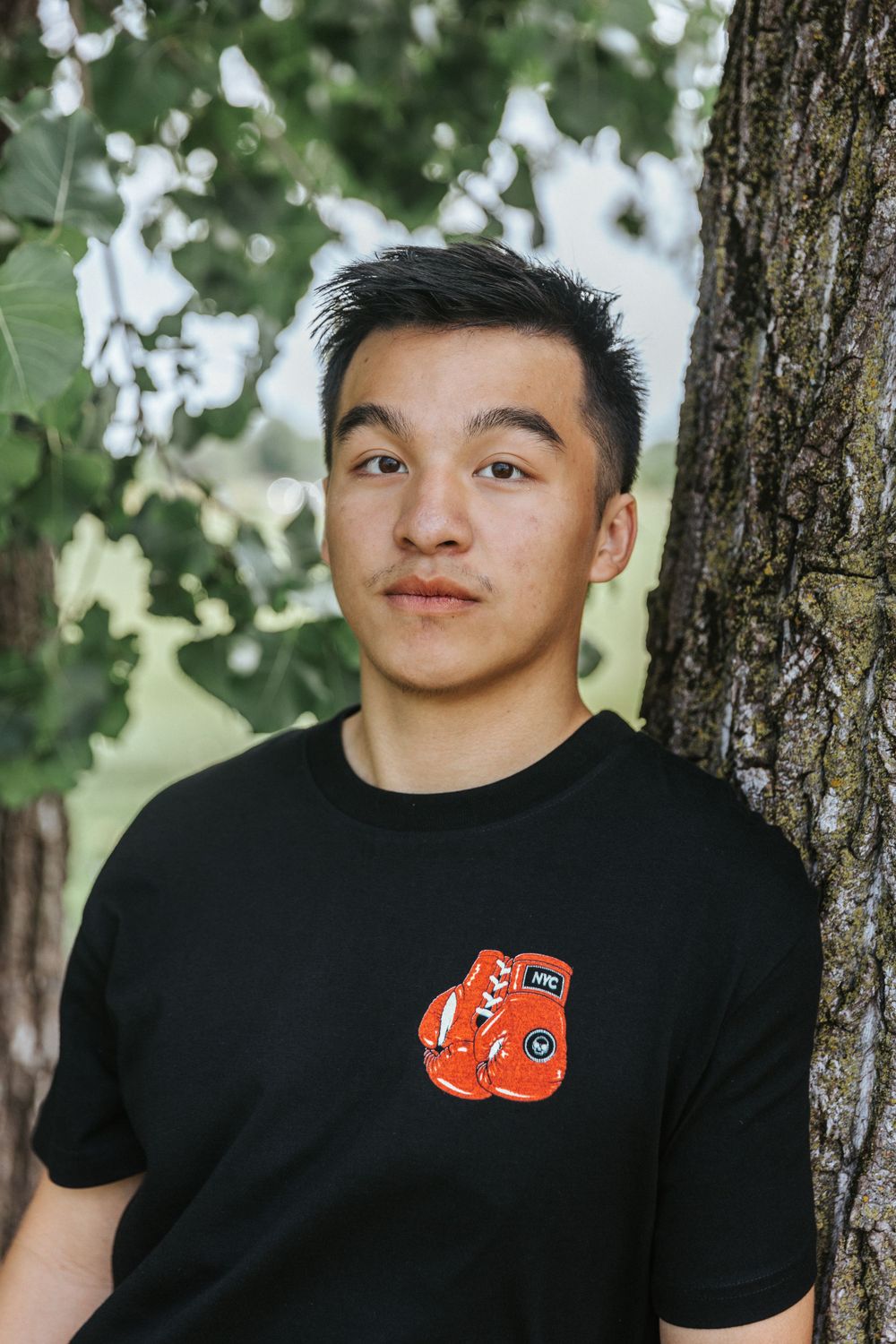 Person in black t-shirt with orange logo poses against tree trunk outdoors.