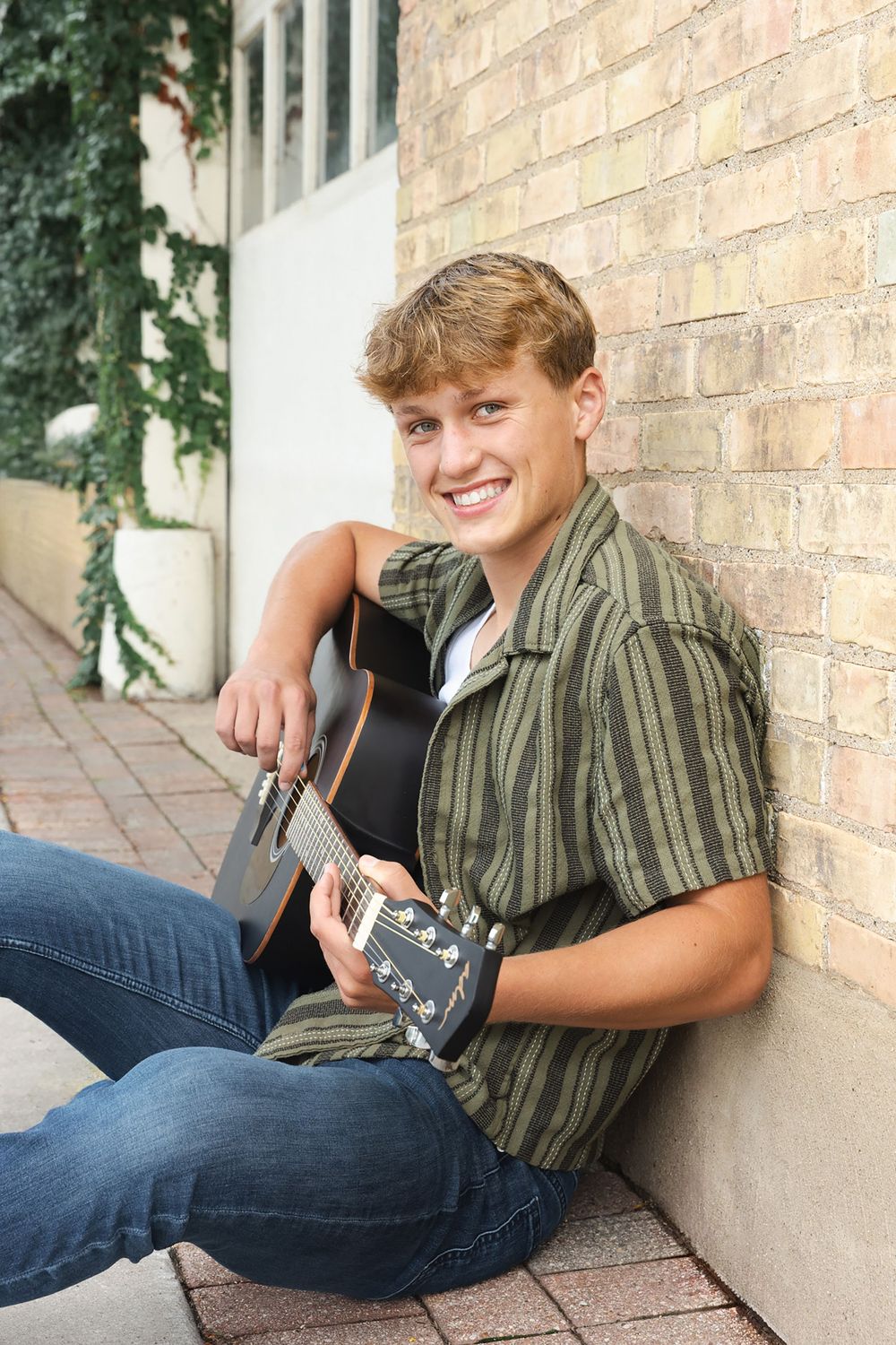 Senior boy smiling at the camera in a downtown setting leaning against brick wall with guitar.
