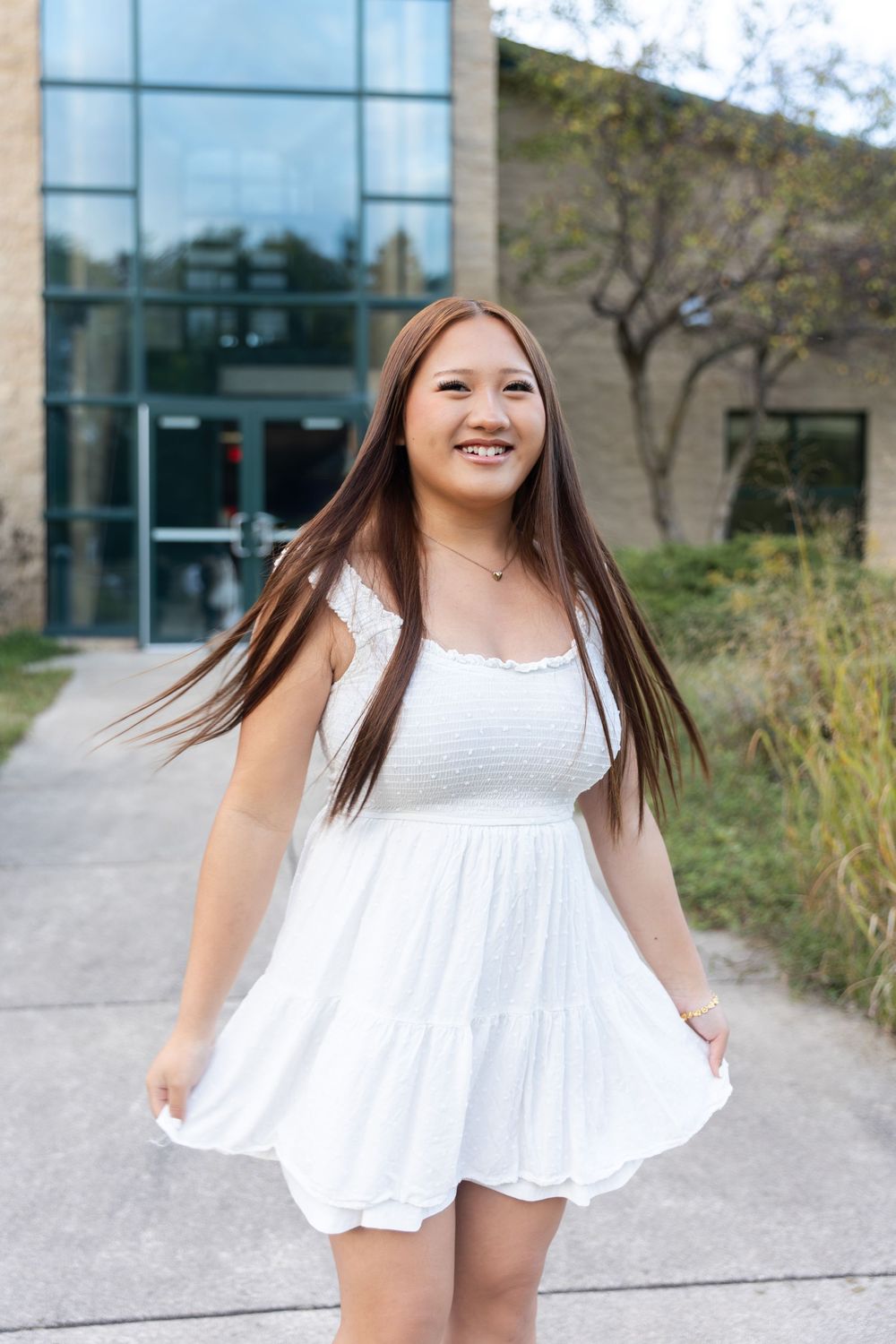 Young woman in white summer dress twirling outdoors near modern building.