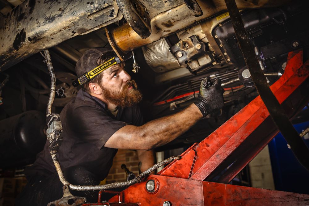 Bearded mechanic wearing headlamp working under a vehicle on a red jack in a repair shop.