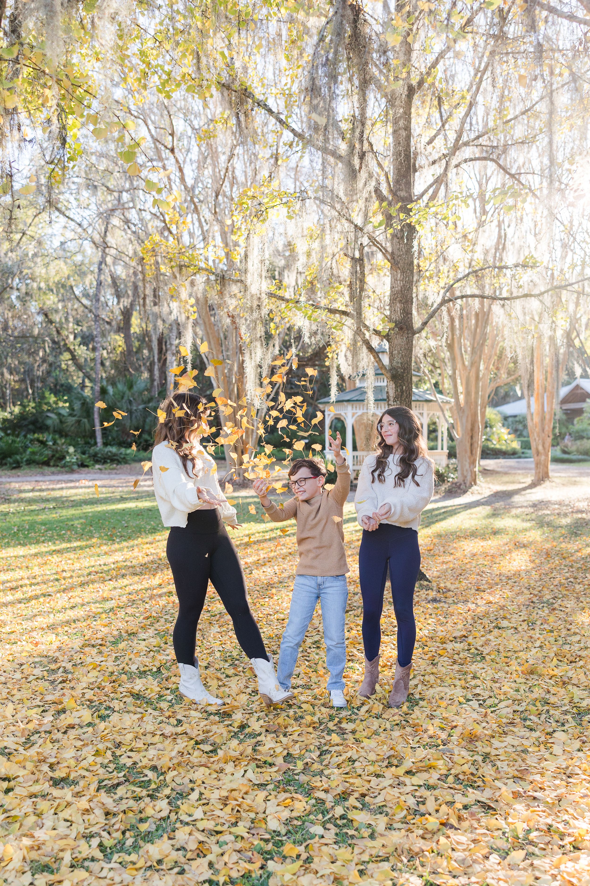 Three people joyfully tossing autumn leaves in a sunny park surrounded by golden foliage and a gazebo.