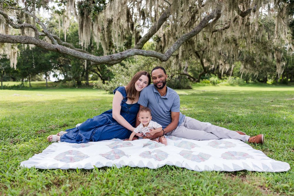 Family of three with parents and baby sitting on a blanket in a lush green park under Spanish moss trees.