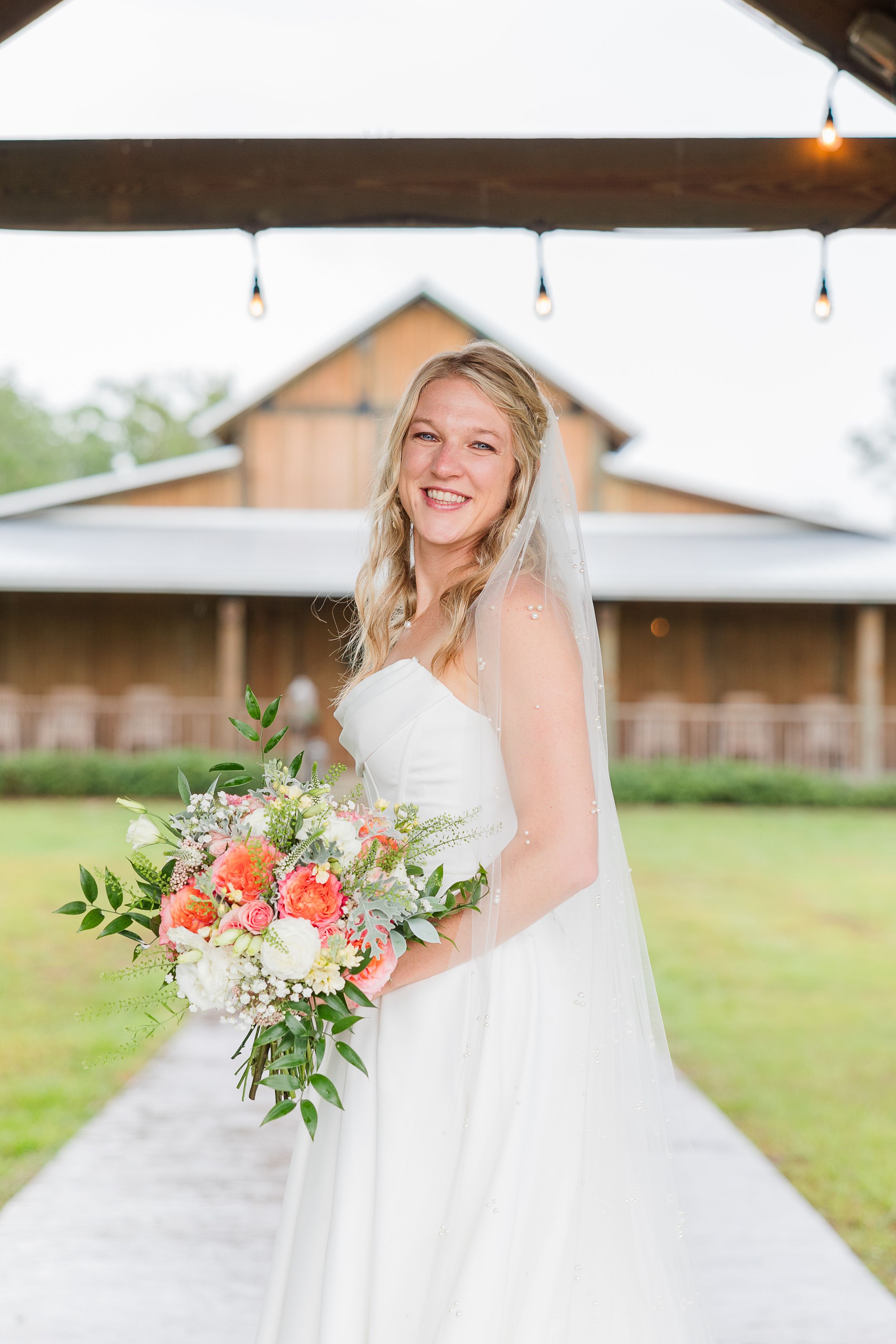 White wedding dress with coral and green bouquet against rustic barn backdrop.