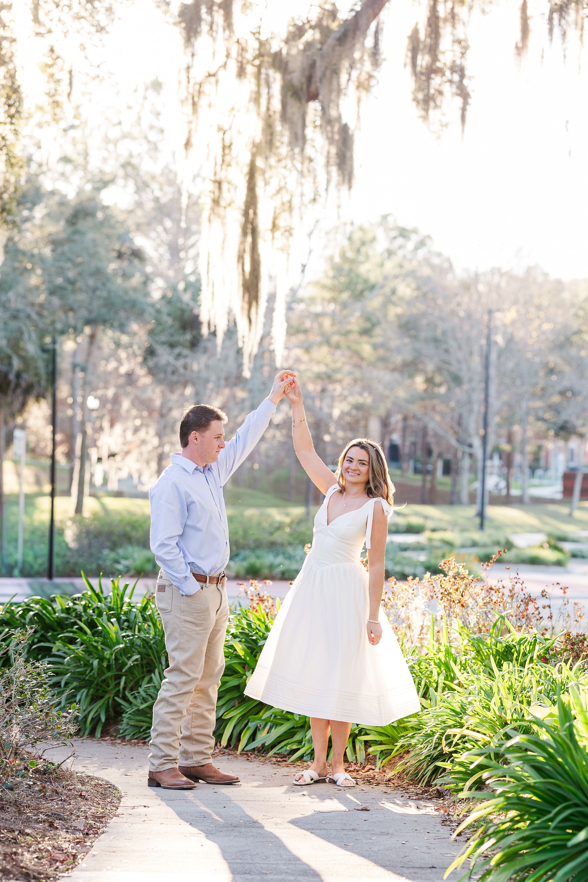 Couple dancing outdoors in a sunlit park with Spanish moss, woman in white dress twirling, lush greenery background.