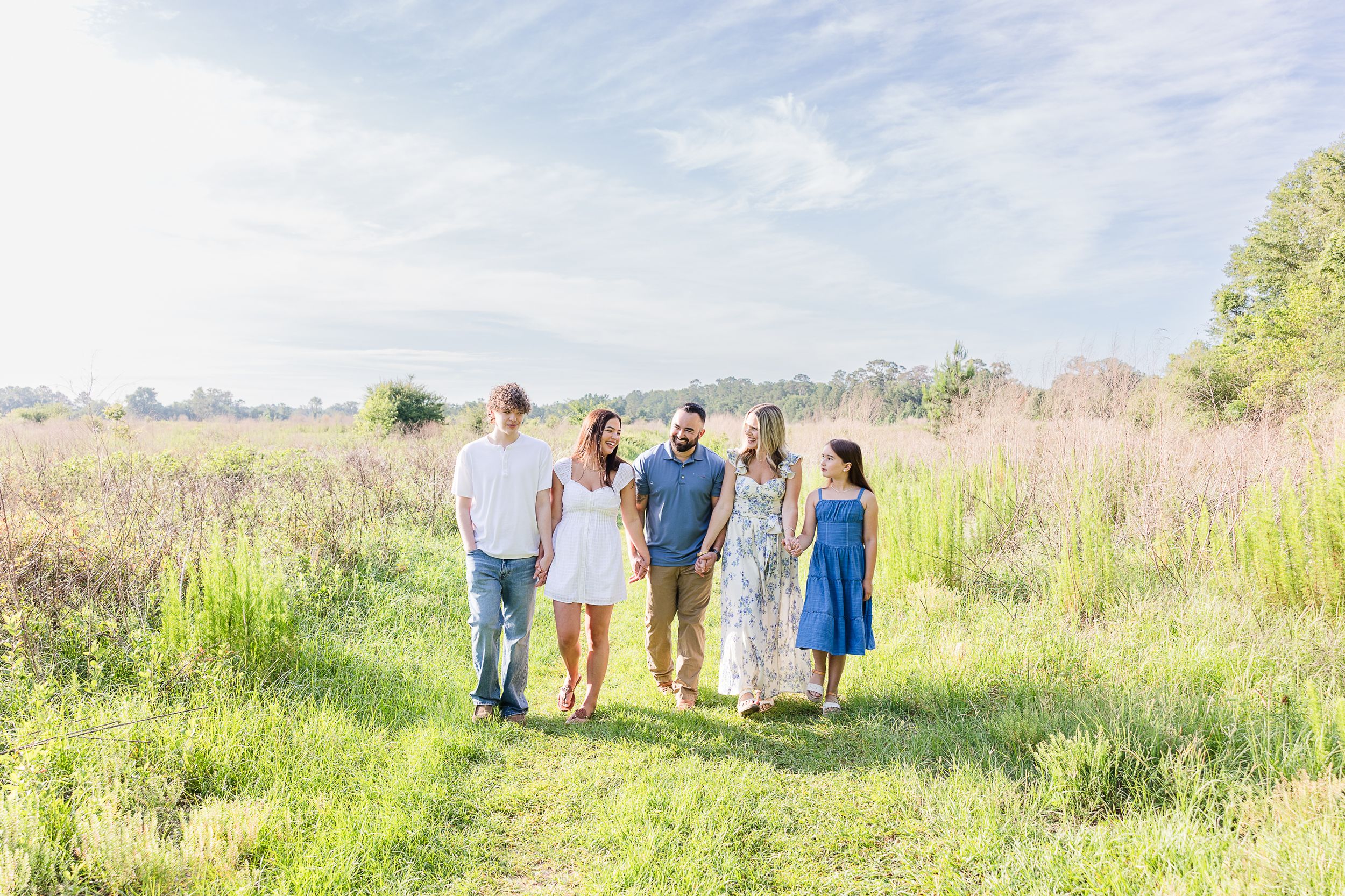 Family of five walking through a sunny wildflower meadow, dressed in white and blue outfits on a bright summer day.