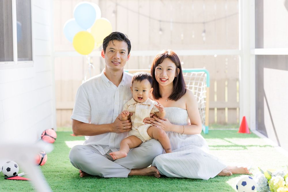 Happy Asian family with baby sitting on green grass with colorful balloons and soccer balls in bright room.