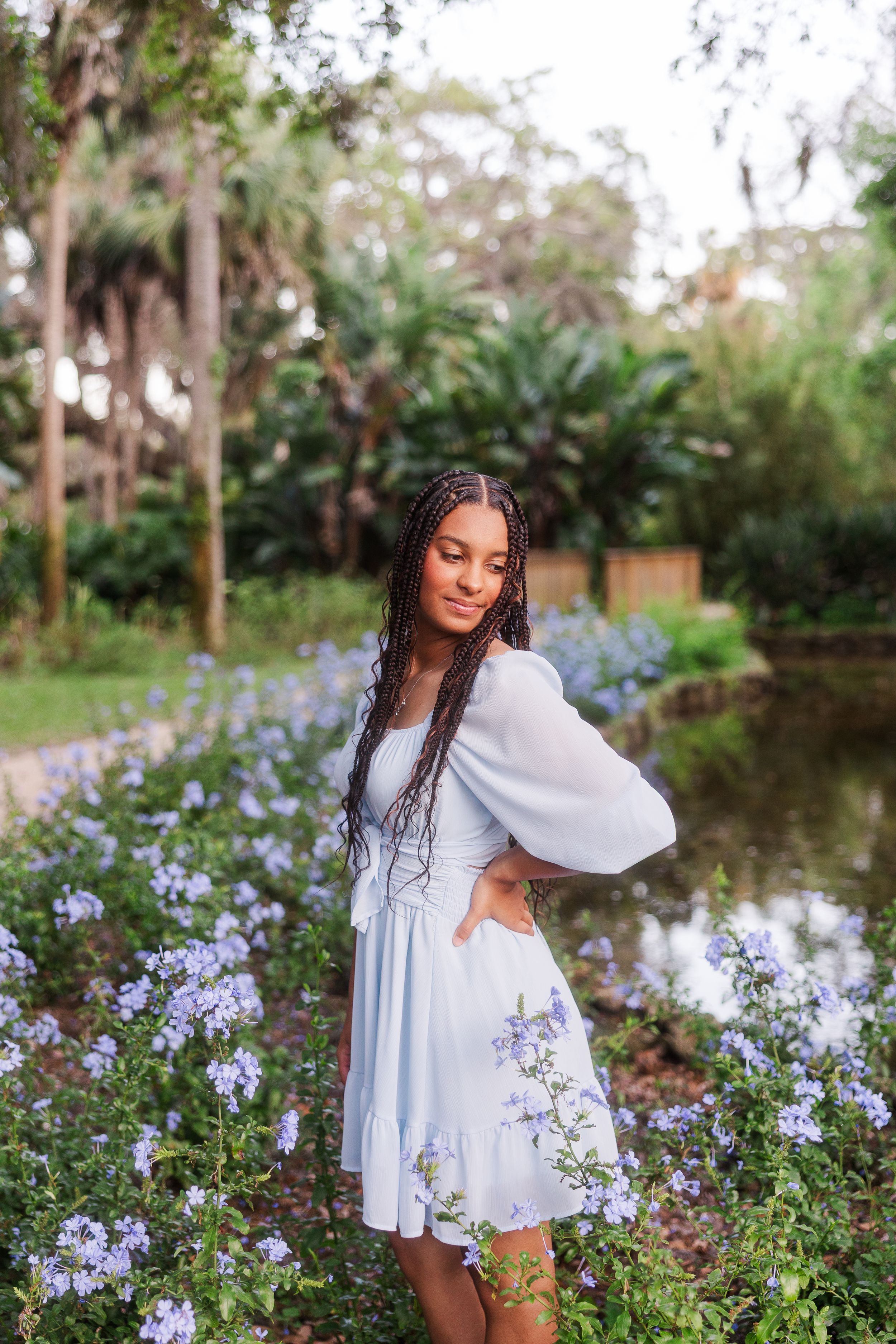 White flowy dress among purple wildflowers near a serene pond with trees in background.