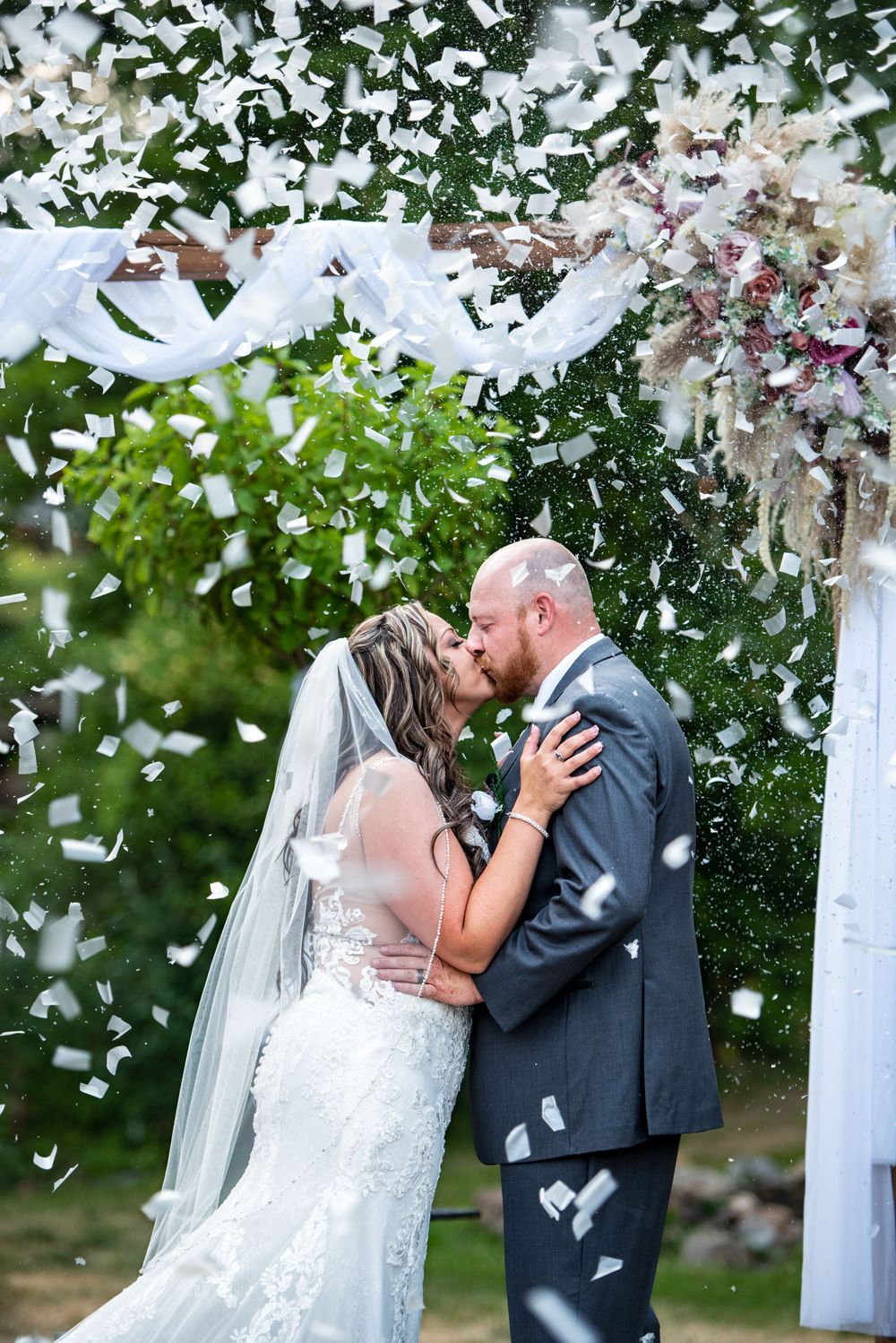 A romantic wedding moment captured under a white arch with confetti falling and floral decorations in an outdoor garden setting.