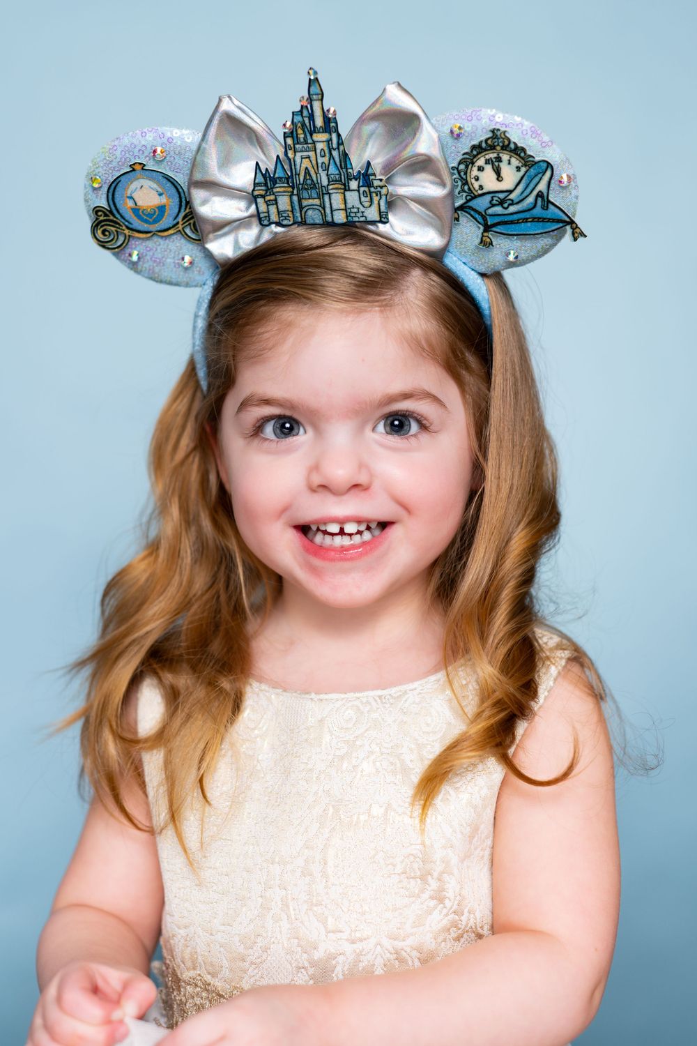 Decorative blue Disney castle themed Mickey ears with silver bow worn with cream colored dress against blue background.