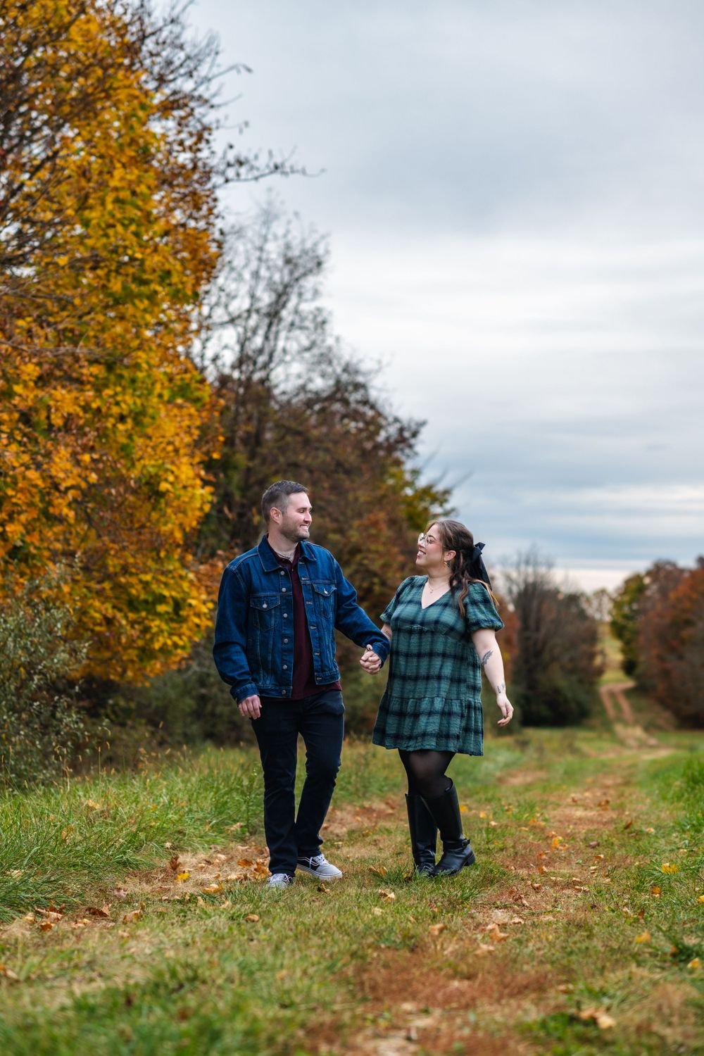 Couple walking together on autumn trail with golden leaves and trees against cloudy sky.