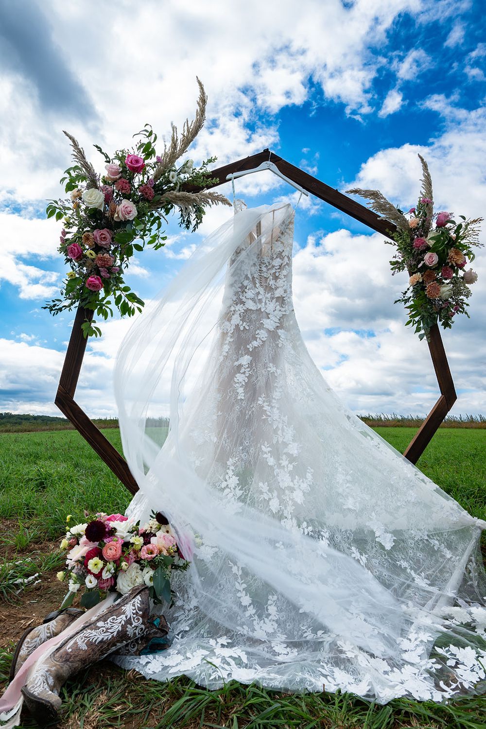 A delicate lace wedding dress hangs on a rustic wooden hexagon arch decorated with pink and white flowers outdoors.