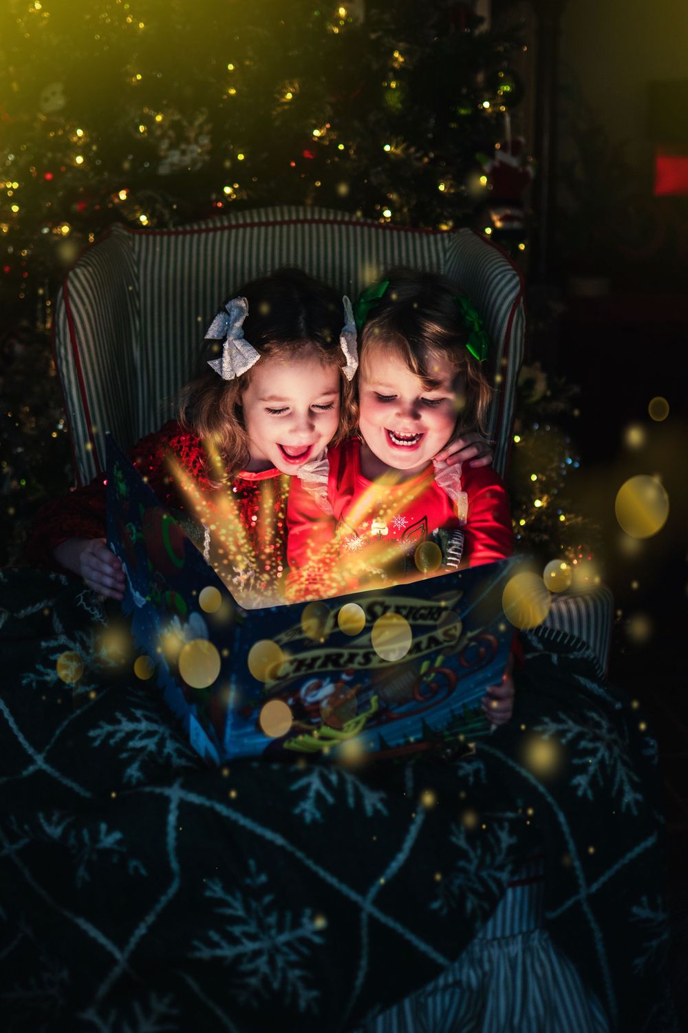 Two children sitting in a chair looking at a glowing book with magical light effects and Christmas tree in background.