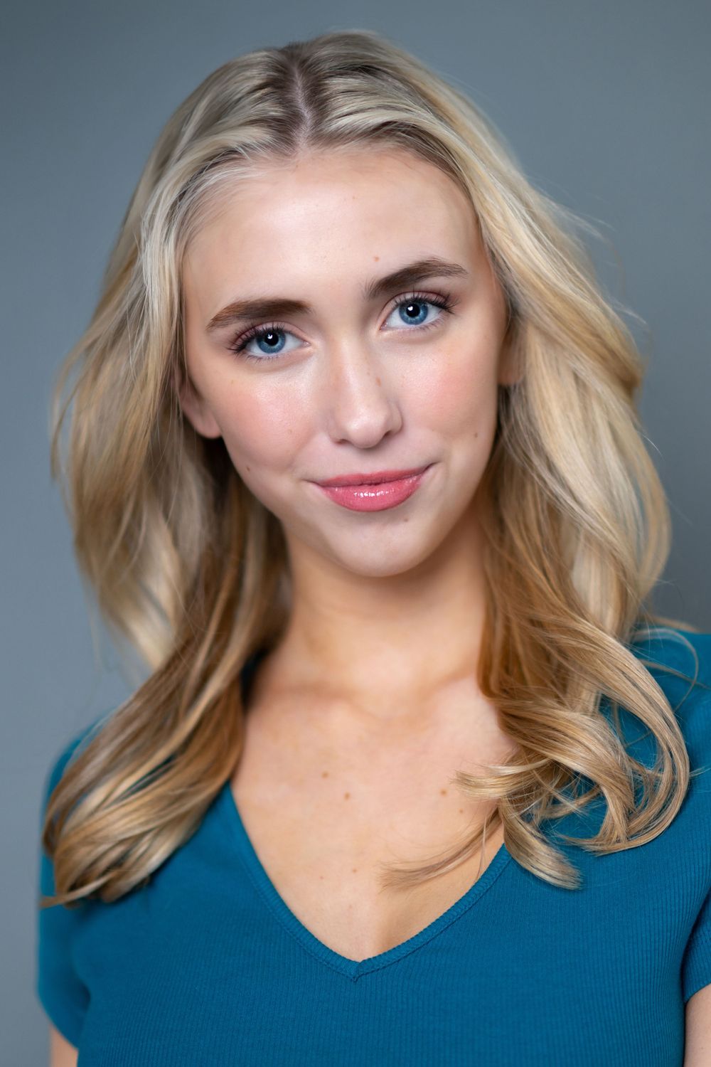 Professional headshot portrait of a person with blonde wavy hair wearing a blue v-neck top against a gray background.