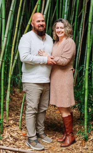 A couple poses together in front of tall green bamboo stalks wearing coordinated neutral outfits.