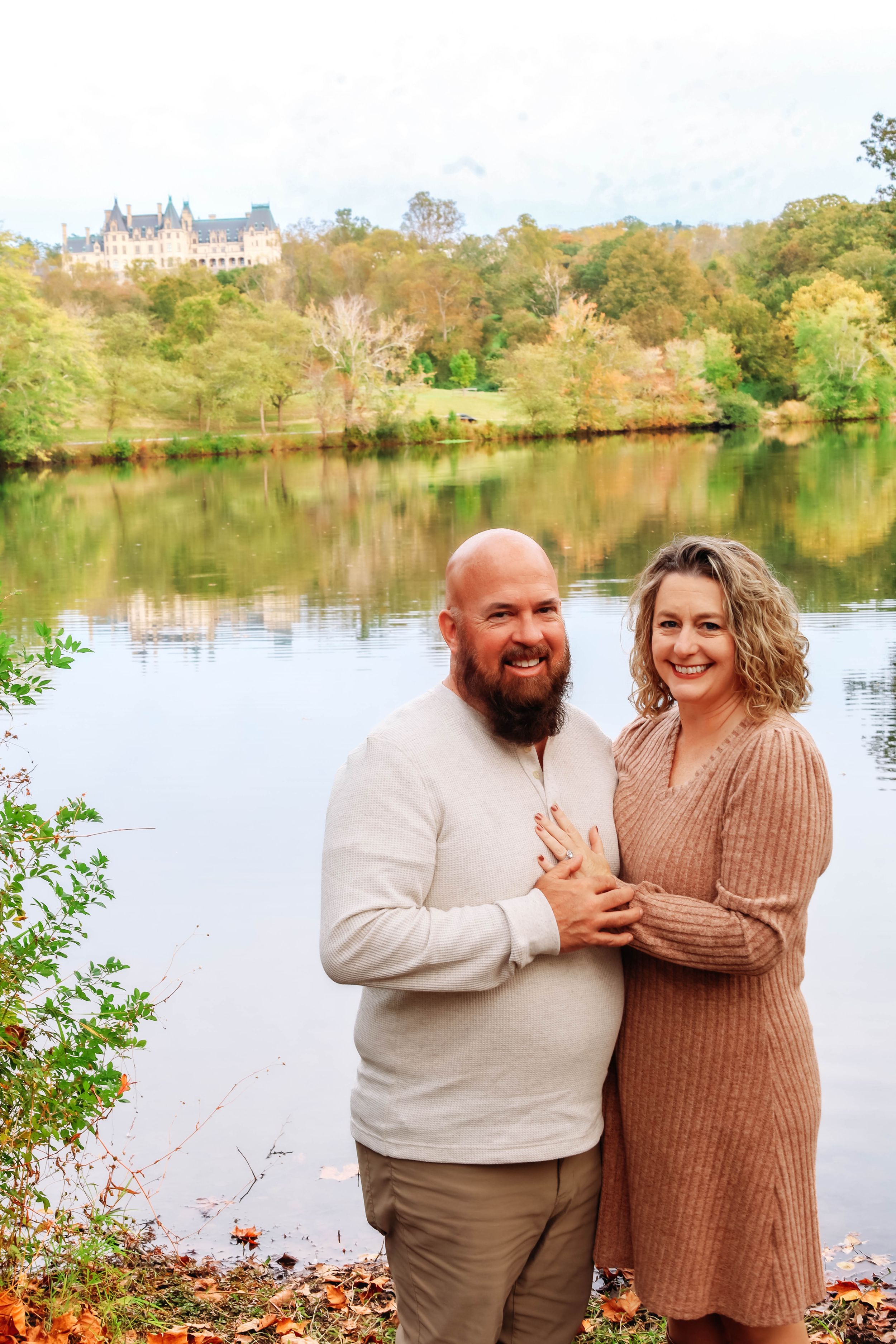 A couple in matching beige sweaters share a romantic moment by a scenic autumn lake.