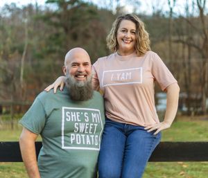 Couple wearing matching t-shirts with sweet potato themed text standing outdoors by a fence.