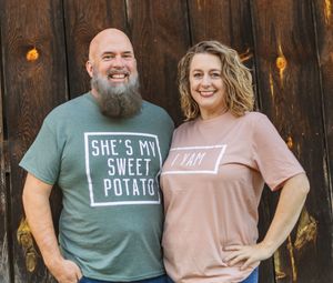 Couple in coordinated t-shirts with sweet potato themed messages posing against wooden wall.