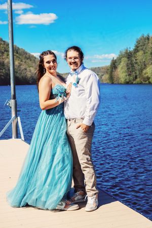 A couple embraces on a dock, with one wearing a flowing turquoise dress against a scenic lake backdrop.