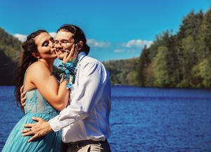A couple shares an intimate moment in formal attire by a blue lake on a sunny day.