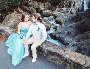 A couple poses together on rocky terrain near a waterfall wearing formal attire.