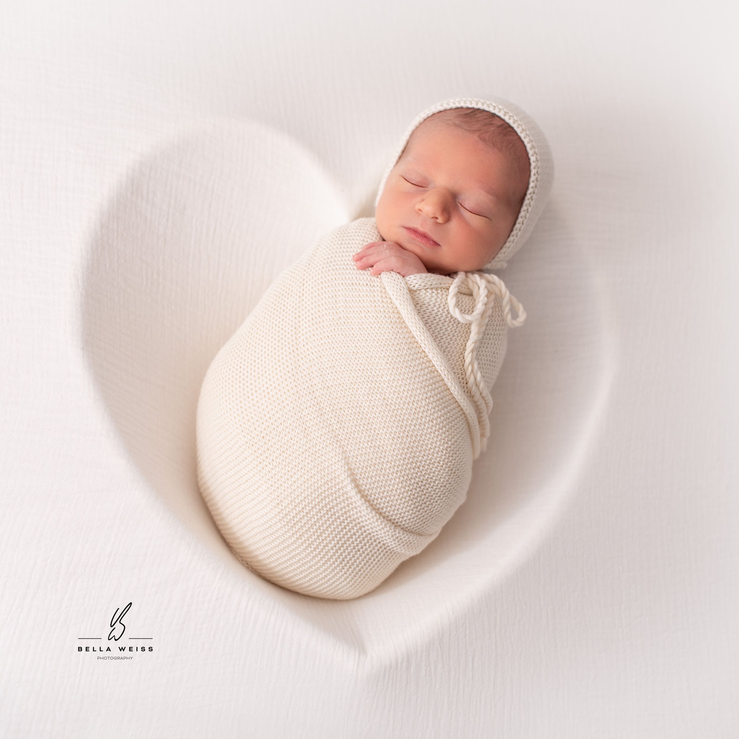 Newborn baby sleeping in white knit wrap on heart-shaped cushion.
