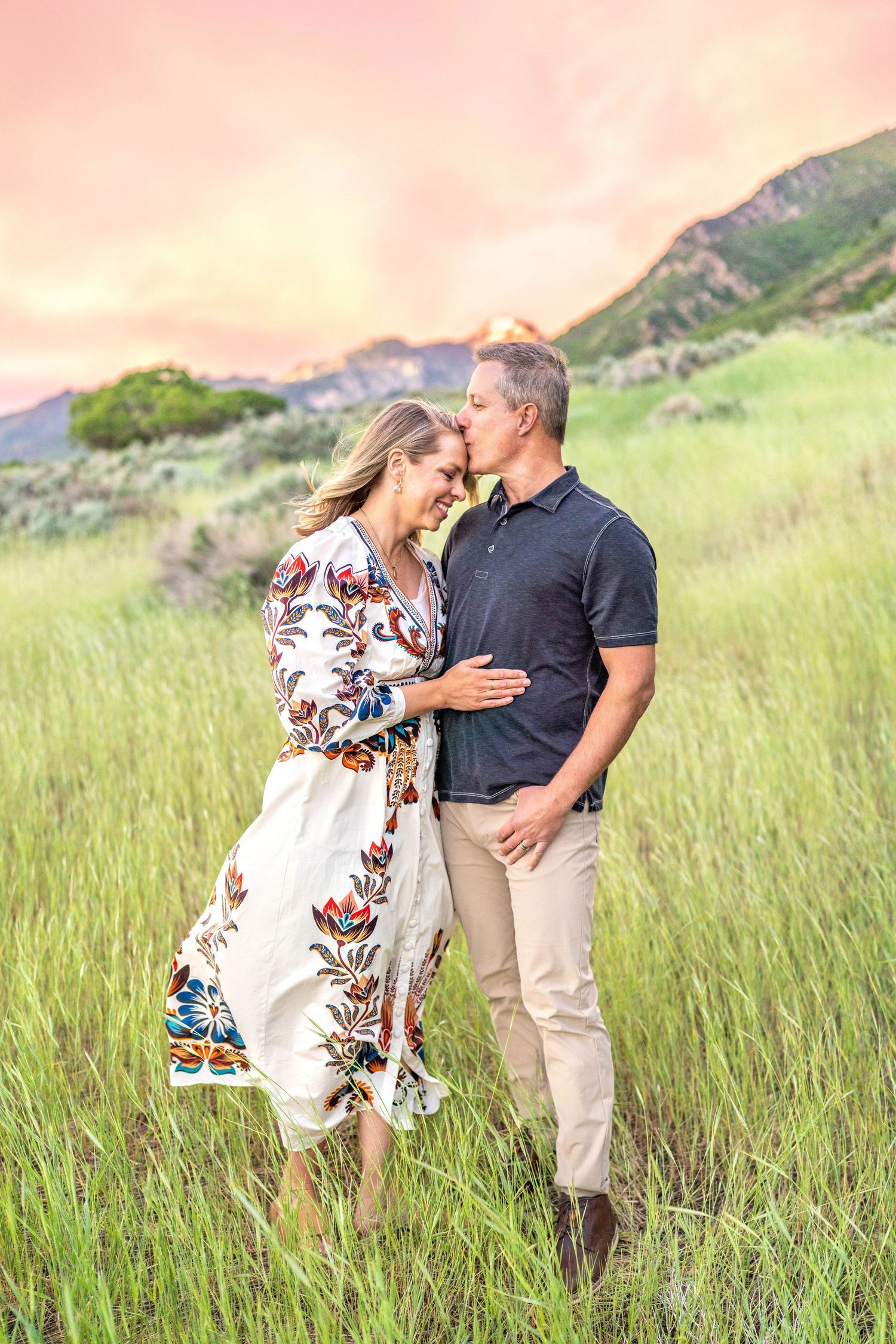 A romantic couple embraces in a grassy meadow with mountains and pink sunset sky in the background at Hidden Valley Park.